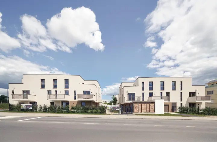 Zwei moderne, helle Wohngebäude mit Balkonen vor blauem Himmel mit weißen Wolken und einer Straße im Vordergrund (Leovita).
