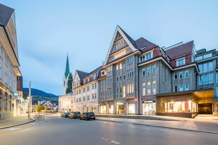 Abendliche Außenansicht des Luger Areals und der Bahnhofstraße mit geparkten Autos und beleuchteten Gebäuden.