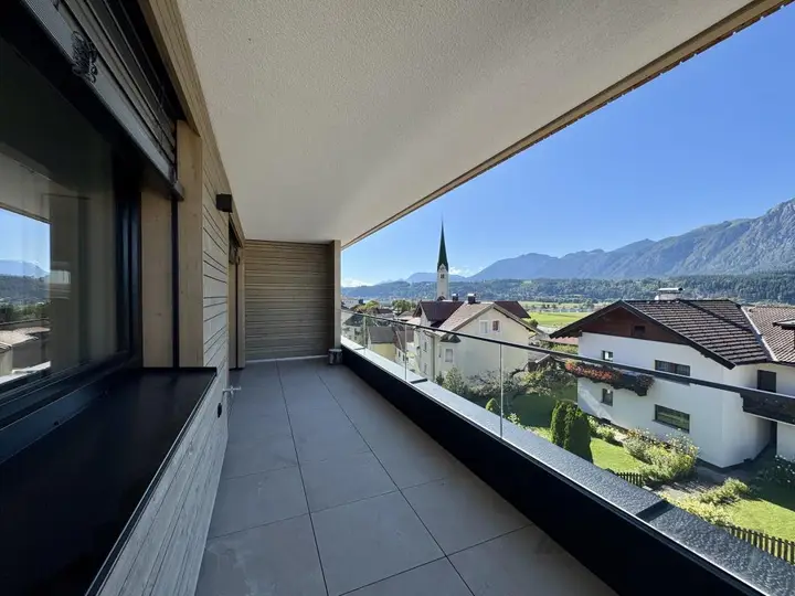 Balkon mit grauen Fliesen, Glasgeländer und weitem Alpenblick bei Sonnenschein.