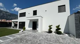 Modernes Einfamilienhaus mit weißer Fassade, gepflegtem Vorgarten und Bergblick unter blauem Himmel.
