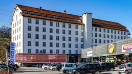 Großes, weißes Gebäude mit vielen Fenstern und einem Parkplatz davor, umgeben von Bergen.