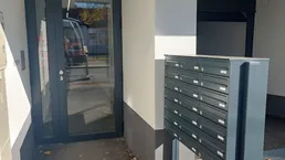 Eingangsbereich eines modernen Gebäudes mit Briefkästen und Glastür an einem sonnigen Tag.