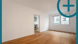 Helles, leeres Zimmer mit Parkettboden und Blick auf die umliegenden Gebäude.