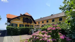 Gelbes Gebäude mit rotem Ziegeldach, grünen Fensterläden und einem kleinen Holzbalkon, umgeben von üppiger Vegetation.