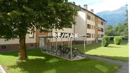 Mehrfamilienhaus mit Fahrradunterstand und gepflegter Grünfläche vor alpiner Kulisse.
