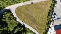 Luftaufnahme eines dreieckigen Grundstücks mit Zufahrtsstraße, umgeben von Wald und einigen Gebäuden.