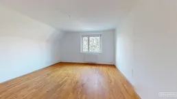 Helles, leeres Zimmer mit Holzparkettboden und einem Fenster, das viel Tageslicht hereinlässt.