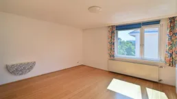 Helles Zimmer mit Holzfußboden, weißen Wänden und einem Fenster mit bunten Vorhängen.