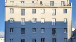Graue Fassade eines Mehrfamilienhauses mit vielen Fenstern und einem Dachbalkon unter blauem Himmel.