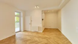 Helles, leeres Zimmer mit Fischgrätparkettboden, Heizkörper und Zugang zu einem Balkon.