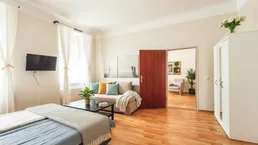 Helles Schlafzimmer mit Parkettboden, Sofa und Zugang zu einem weiteren Raum.