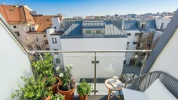 Heller Balkon mit Stadtblick, Pflanzen und gemütlicher Sitzgelegenheit unter blauem Himmel.