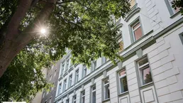 Historische Fassade eines Mehrfamilienhauses mit grünen Akzenten und großen Fenstern unter einem Baum.
