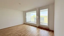 Helles, leeres Zimmer mit Parkettboden und großen Fenstern, die Zugang zu einem Balkon bieten.