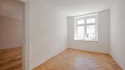 Helles Zimmer mit Fischgrätparkettboden und einem Fenster, das in einen weiteren Raum führt.