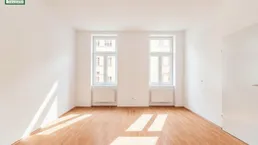 Helles, leeres Zimmer mit Parkettboden und zwei Fenstern, das viel Tageslicht hereinlässt.