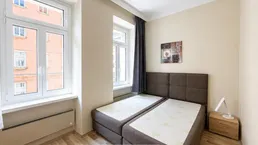 Helles Schlafzimmer mit zwei Einzelbetten, Nachttisch, Laminatboden und großen Fenstern.