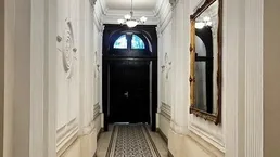 Eleganter Flur mit hohen Decken, Stuckverzierungen und einem gemusterten Läufer, der zu einer dunklen Holztür führt.