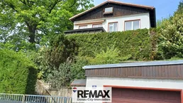 Weißes Haus mit dunklem Dach, umgeben von üppigem Grün und hohen Bäumen.