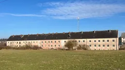 Langes Wohngebäude mit vielen Fenstern und einem weiten Feld im Vordergrund unter blauem Himmel.