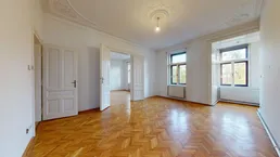 Geräumiges Wohnzimmer mit Fischgrätparkett, hohen Decken und Stuckverzierungen, hell durch große Fenster.