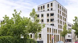 Modernes Mehrfamilienhaus mit heller Ziegelfassade und markanten goldenen Fensterrahmen an einer städtischen Straße.