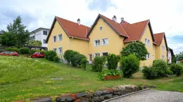 Gelbes Wohngebäude mit rotem Ziegeldach und grüner Gartenanlage unter bewölktem Himmel.