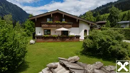 Traditionelles Alpenhaus mit gepflegtem Garten, Terrasse und Bergblick unter blauem Himmel.