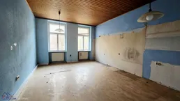 Leerer Raum in Renovierungszustand mit blauen Wänden, Holzdecke und zwei großen Fenstern.