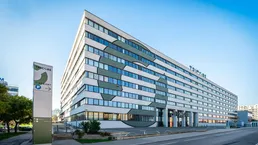 Modernes Bürogebäude mit markanter Fassadengestaltung und Parkplatzzufahrt unter blauem Himmel.