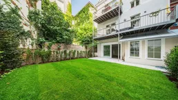 Grüner Rasen und eine Terrasse vor einem Gebäude mit Balkonen und bewachsenen Wänden.