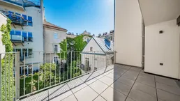 Heller Balkon mit Fliesenboden und Blick auf begrünte Innenhöfe und umliegende Gebäude.