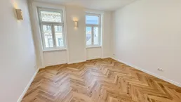 Helles Zimmer mit elegantem Fischgrätparkett und zwei großen Fenstern, ideal als Wohn- oder Schlafzimmer.