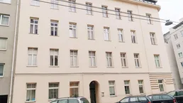 Die Fassade eines Mehrfamilienhauses mit vielen Fenstern und parkenden Autos auf der Straße.