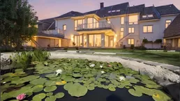 Großzügiges Anwesen mit Teich und beleuchteter Fassade bei Sonnenuntergang, umgeben von Grünflächen.