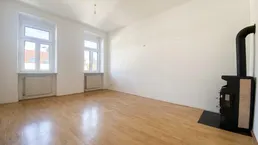 Helles Zimmer mit Holzboden, zwei Fenstern und einem gemütlichen Holzofen als Wärmequelle.