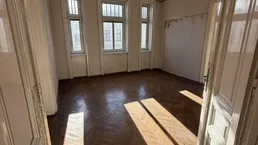 Helles Zimmer mit Fischgrätparkett und hohen Fenstern, durch die Sonnenlicht einfällt.