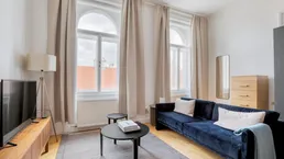 Helles Wohnzimmer mit blauem Sofa, Holzboden und hohen Bogenfenstern, die viel Tageslicht hereinlassen.