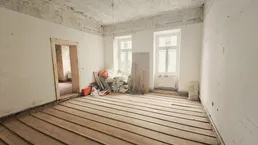 Leerer Raum in Renovierung mit freigelegten Holzdielen, hohen Decken und großen Fenstern.