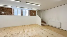 Großer, leerer Raum mit unverputzten Ziegelwänden, Sperrholzfußboden und vielen Deckenleuchten.