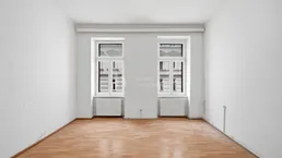 Helles Zimmer mit zwei Fenstern und Holzboden, bereit zur individuellen Gestaltung.