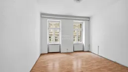 Helles, leeres Zimmer mit Laminatboden und zwei Fenstern, die auf die Straße blicken.