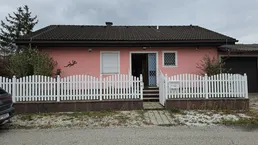 Einfamilienhaus mit rosafarbener Fassade, weißem Lattenzaun und Vorgarten unter bewölktem Himmel.