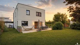 Modernes Einfamilienhaus mit großer Rasenfläche und möblierter Terrasse im Sonnenuntergang.