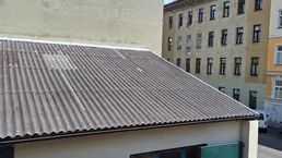 Ansicht eines Nebengebäudes mit Wellblechdach und grüner Tür, umgeben von höheren Gebäuden unter blauem Himmel.