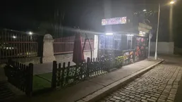 Nachtaufnahme eines Imbissstandes am Straßenrand mit kleiner eingezäunter Fläche und rotem Sonnenschirm.