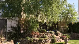 Gepflegter Gartenbereich mit Rasenfläche, Bäumen und terrassierten Blumenbeeten unter blauem Himmel.