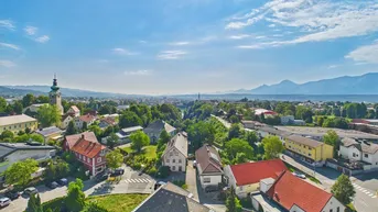 Panoramablick über eine Stadt mit Häusern, viel Grün und Bergen im Hintergrund unter blauem Himmel.