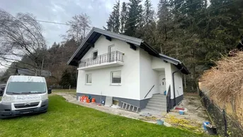 Modernes Einfamilienhaus mit weißer Fassade, dunklem Dach und kleinem Balkon, umgeben von Grünflächen.