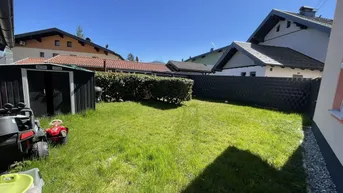 Gepflegter Gartenbereich mit Rasenfläche, Hecke und Gartenhaus unter blauem Himmel.